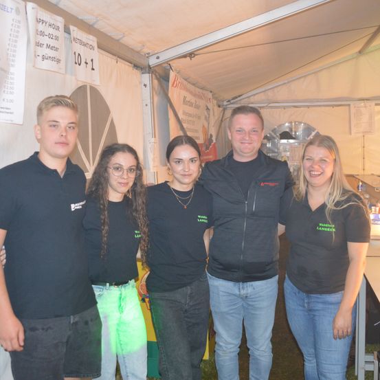 Fünf Personen in gleichen schwarzen T-Shirts posieren für ein Foto in einem Zelt. Das Zelt hat ein weißes Dach und einen Banner mit Text. Der Hintergrund zeigt eine weiße Wand mit Preisschildern.