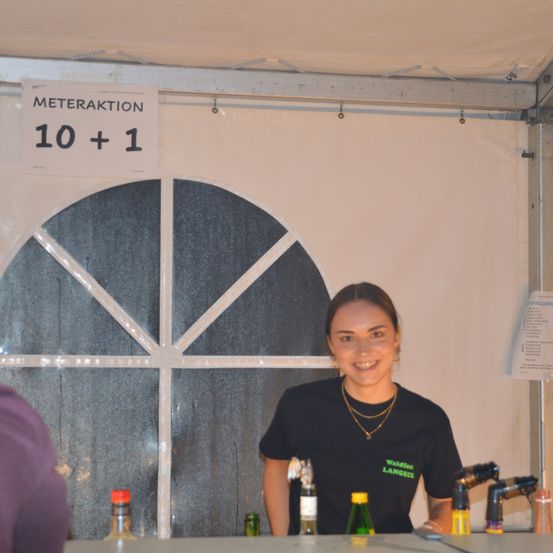 Eine Frau steht hinter einem Tisch mit Flaschen und einem Mikrofon und lächelt vor einem Glasfenster mit einem Schild, das 'Meteraktion 10 + 1' steht.