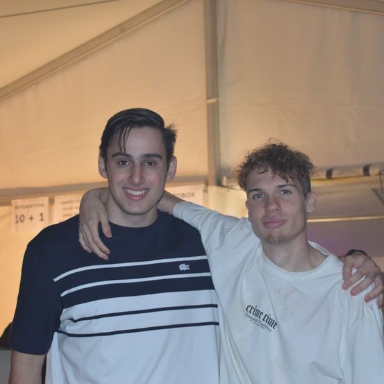 Zwei Männer lächeln und posieren für ein Foto. Der Mann auf der linken Seite trägt ein blau-weiß gestreiftes Shirt. Der Mann auf der rechten Seite trägt ein weißes T-Shirt mit den Worten 'crime time' darauf. Sie stehen beide unter einem Zelt.