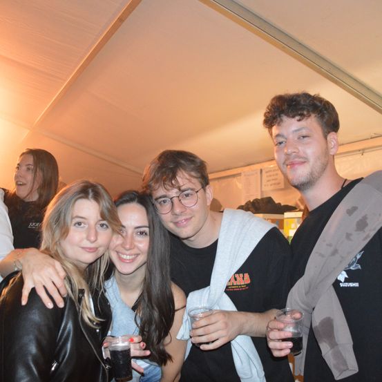 Eine Gruppe von Freunden lächelt und posiert für ein Foto auf einer Party. Sie halten Getränke in Gläsern. Ein Mann mit Brille und einer weißen Kapuzenjacke steht in der Mitte, und zwei Frauen sind an den Seiten. Der Hintergrund ist verschwommen mit anderen Leuten und einer weißen Decke.