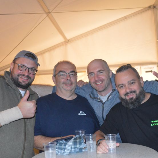 Vier Männer sitzen an einem Tisch mit Gläsern, zwei von ihnen zeigen den Daumen nach oben. Der Mann auf der rechten Seite hat einen Bart und trägt ein Shirt mit den Worten 'Waldfang Lang'. Sie alle lächeln und scheinen für ein Foto zu posieren.