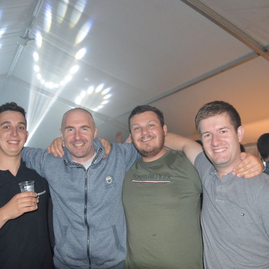 Vier Männer stehen zusammen in einem Zelt mit weißer Decke und lächeln für ein Foto. Ein Mann hält ein Glas.