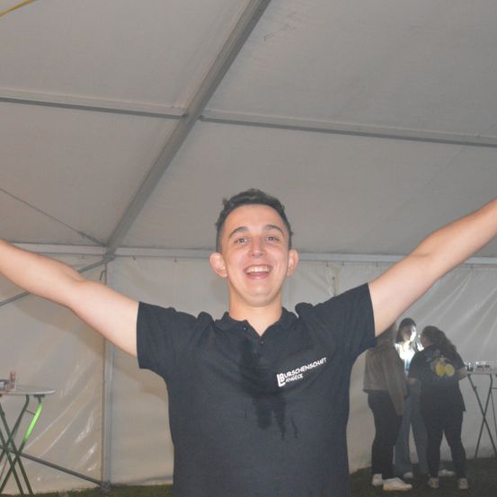 Ein Mann in einem schwarzen T-Shirt steht in einem Zelt, Arme ausgestreckt und lächelt. Hinter ihm steht ein Tisch mit Flaschen, und mehrere Leute stehen in der Nähe, einer trägt ein gelbes Shirt.