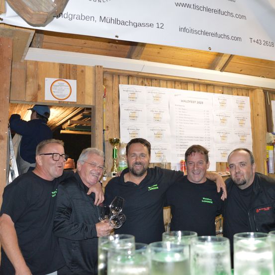 Fünf Männer posieren für ein Foto an einem Tisch mit Weingläsern. Sie tragen schwarze Hemden. Hinter ihnen ist eine Wand mit einem Plakat. Ein Mann steht auf einer Leiter in der Nähe des Eingangs.