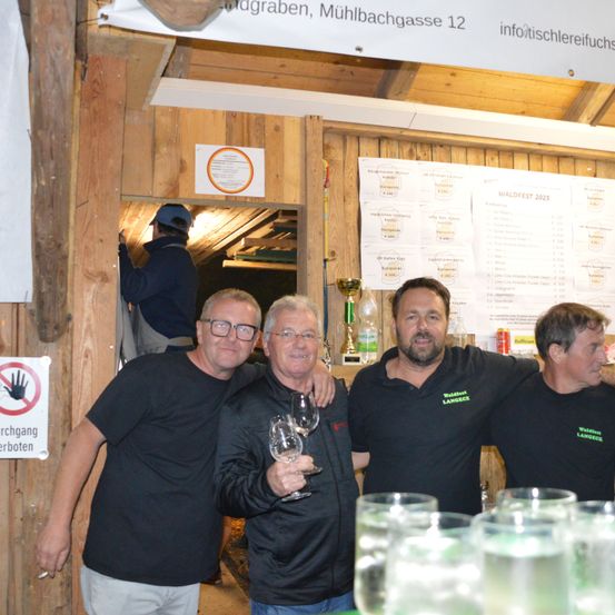 Vier Männer in schwarzen T-Shirts, einer hält ein Weinglas, stehen vor einer Holzkabine mit einem Pokal. Ein Schild an der Wand sagt 'Handverbot'. Ein Mann in einem blauen Hut ist im Hintergrund.
