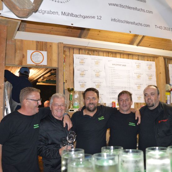 Fünf Männer in schwarzen T-Shirts stehen in einer rustikalen Umgebung zusammen. Einer hält einen Pokal. Hinter ihnen ist ein Banner mit Veranstaltungsdetails an einer Holzwand angebracht.