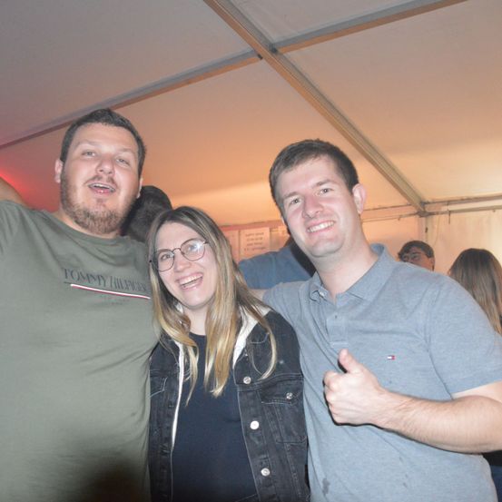 Drei Menschen lächeln und posieren für ein Foto in einer sozialen Umgebung. Der Mann auf der linken Seite trägt ein Tommy Hilfiger-Shirt. Die Frau in der Mitte trägt eine Brille und eine Jacke. Der Mann auf der rechten Seite gibt einen Daumen hoch.