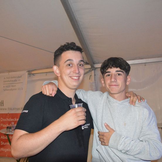 Zwei junge Männer posieren für ein Foto unter einem Zelt. Ein Mann in einem schwarzen Shirt hält einen Becher, während der andere in einem grauen Hoodie seinen Arm um seine Schulter hat. Ein Banner hinter ihnen hat deutschen Text.