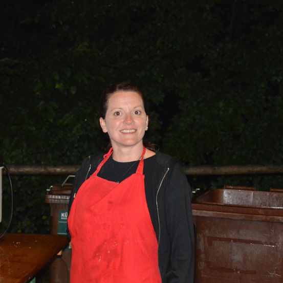 Eine Frau in einem roten Schürze steht nachts draußen, lächelt und hat einen Holztisch und Mülleimer im Hintergrund.