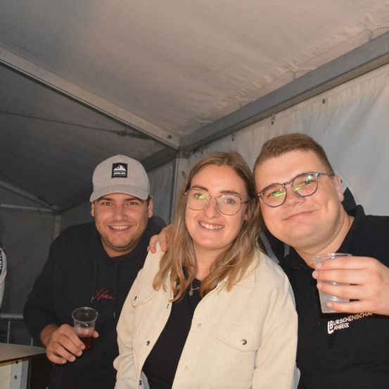 Drei Personen, zwei Männer und eine Frau, posieren für ein Foto unter einem Zelt. Sie lächeln und halten Gläser. Die Frau trägt eine Brille und eine Jacke. Der Mann rechts trägt ein Poloshirt. Der Mann links trägt eine Kappe. Der Hintergrund ist weiß.