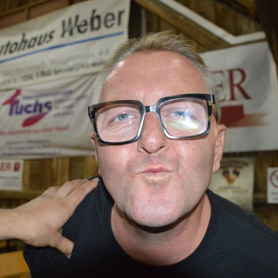 Ein Mann mit schwarzen Brille und einem schwarzen T-Shirt posiert für ein Foto vor einem Banner, das 'Tuchos' sagt.