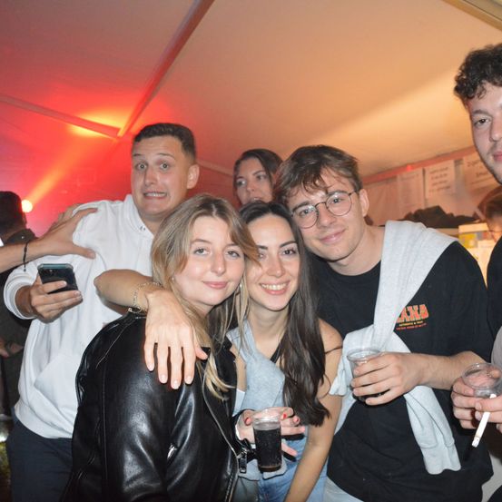 Eine Gruppe von Freunden auf einer Party, die für ein Foto posieren. Der Mann in Weiß hält ein Handy, während andere Getränke und eine Zigarette haben.