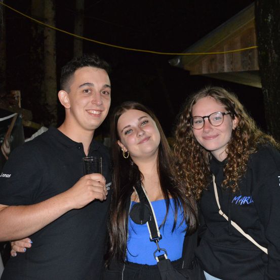 Drei Menschen stehen nachts zusammen. Ein Mann in einem schwarzen Shirt lächelt, eine Frau mit einem blauen Top und eine Frau mit Brille. Sie stehen draußen in der Nähe einer Holzstruktur.