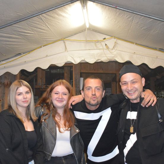 Vier Personen stehen unter einem Zelt. Zwei Männer tragen schwarze und weiße T-Shirts mit einem großen Buchstaben Z. Die Frauen stehen auf der linken und rechten Seite.