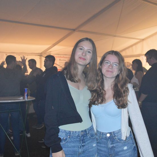 Zwei Frauen stehen nebeneinander in einem Zelt bei einer Veranstaltung, lächeln und posieren für ein Foto. Im Hintergrund sind andere Menschen, einige stehend und andere sitzend, und ein Tisch mit einer Dose darauf.