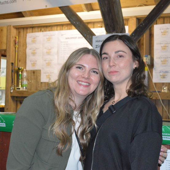 Zwei Frauen stehen lächelnd nebeneinander vor einer Holzwand. Die Frau auf der linken Seite hat lange blonde Haare und trägt eine grüne Jacke. Die Frau auf der rechten Seite hat dunkle Haare und trägt eine schwarze Jacke. Hinter ihnen sind mehrere Papiere an der Wand angeheftet und ein grüner Container.