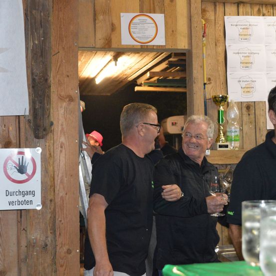 Zwei Männer in schwarzen Shirts stehen nebeneinander, lächeln, mit einem Pokal und einem Weinglas. Ein Schild an der Wand sagt 'Durchgang verboten'. Dahinter klettert eine Person mit rotem Hut eine Leiter hoch.