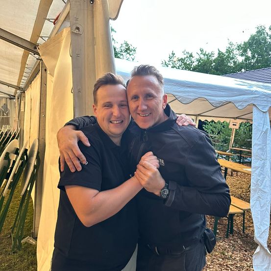 Zwei Männer in identischen schwarzen Jacken und Hosen stehen unter einem weißen Zelt, lächeln und posieren für ein Foto.