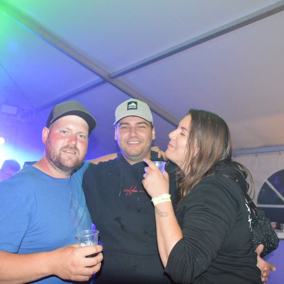 Drei Menschen lächeln und posieren für ein Foto. Zwei Männer und eine Frau, unter einem Zelt mit farbigen Lichtern. Die Frau hält einen Becher.