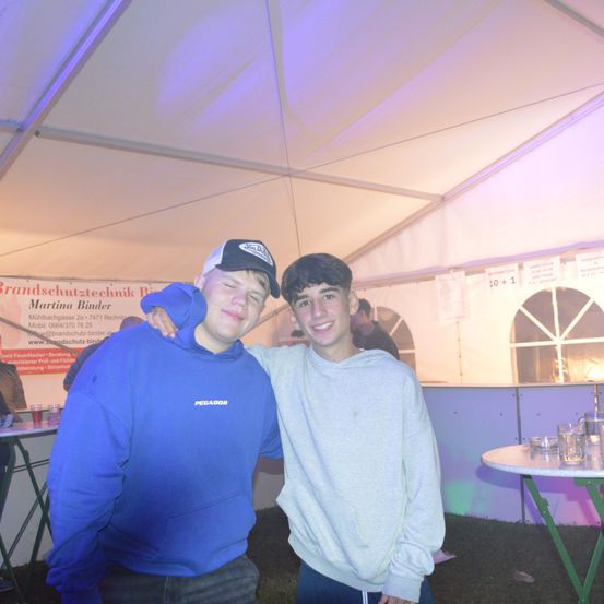 Zwei junge Männer stehen in einem Zelt nebeneinander, einer trägt eine blaue Kapuzenjacke und der andere ein graues Sweatshirt. Sie lächeln und scheinen für ein Foto zu posieren. Dahinter steht ein weißes Zelt mit Schildern und einem Schreibtisch.