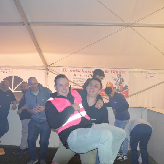 Eine Gruppe von Menschen steht in einem Zelt. Zwei Männer sitzen auf den Schultern des anderen. Ein Banner im Hintergrund trägt die Aufschrift 'Brandschutz mit Binder'.