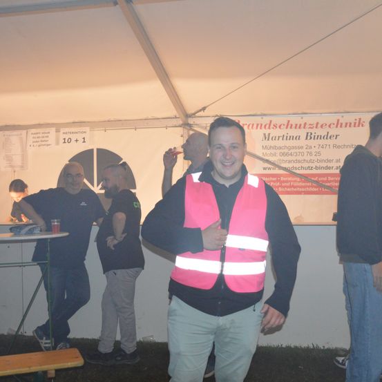 Ein Mann in einer pinken Sicherheitsweste posiert für ein Foto in einem Zelt, umgeben von anderen Leuten. Hinter ihm befindet sich ein Banner mit Text und Zahlen.