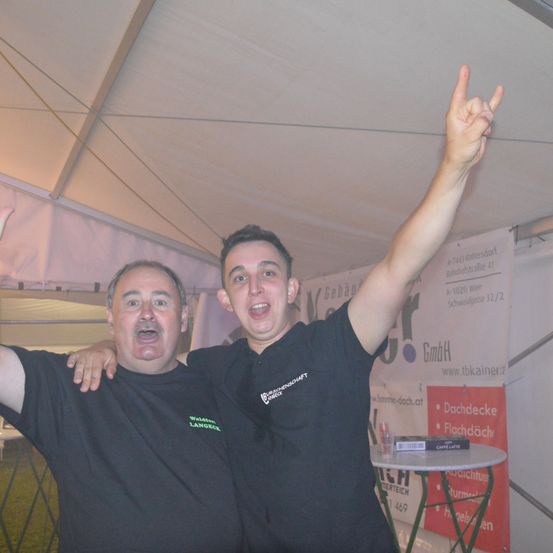Zwei Männer stehen in einem Zelt, lächeln und posieren für ein Foto. Ein Mann macht ein Rock-Zeichen mit der Hand, während der andere Mann seine Schulter hält. Es gibt einen kleinen Tisch mit einem Glas und einer Schachtel darauf. Hinter ihnen ist eine Wand mit einem Schild.
