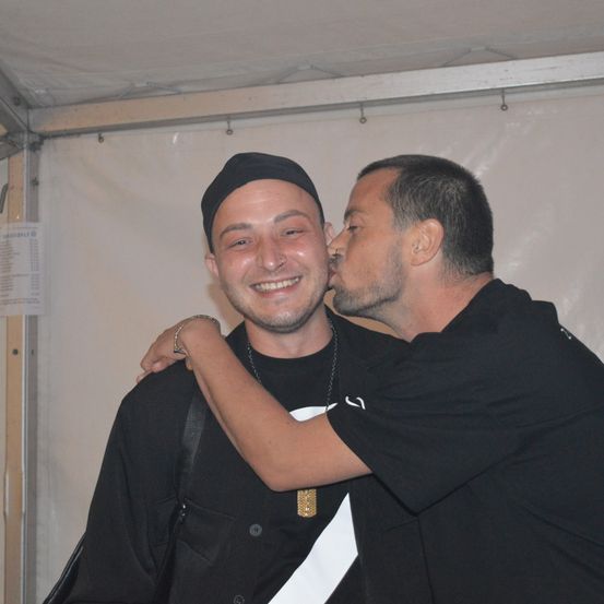 Zwei Männer in schwarzen T-Shirts, einer mit einer schwarzen Kappe, umarmen sich und küssen sich in einem Zelt.