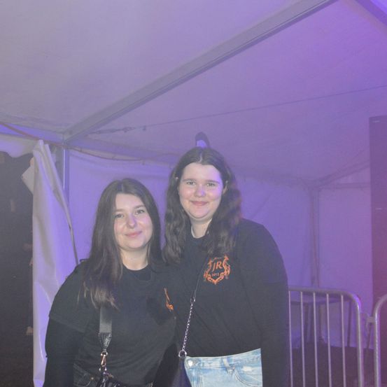 Zwei junge Frauen stehen unter einem lila Licht und lächeln. Sie tragen schwarze T-Shirts, eines mit einem Logo, und stehen in der Nähe einer weißen Leinwand und eines Metallzauns.