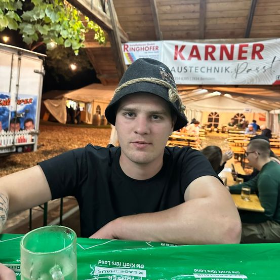 Ein Mann mit Hut und schwarzem T-Shirt sitzt in einem Biergarten. Über ihm hängt ein Banner mit der Aufschrift 'Ringhofer' und ein weiteres mit 'Karner'. Auf dem Tisch steht ein Glas mit grüner Flüssigkeit.