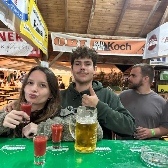 Drei Personen posieren für ein Foto bei einer Outdoor-Veranstaltung. Der Mann in der Mitte gibt einen Daumen hoch, während die Frau links von ihm ein Getränk hält. Sie sitzen an einem Tisch mit Bier und Getränken davor. Über ihnen hängen Banner und Schilder.