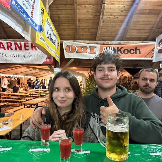 Zwei Personen sitzen vor einem Tisch mit Getränken bei einer Outdoor-Veranstaltung. Ein Mann gibt einen Daumen hoch und eine Frau lächelt. Banner und Menschen sind im Hintergrund.