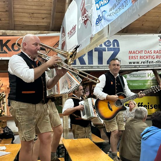 Eine Band spielt in einem Raum mit Holzbänken und Tischen. Ein Mann spielt Trompete, ein anderer Gitarre und ein Mann Akkordeon. Es sitzen Leute auf den Bänken und eine Fahne hängt an der Wand.
