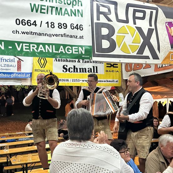 Eine Gruppe von Musikern spielt in einer Outdoor-Umgebung. Sie sind in traditioneller Kleidung gekleidet, einer spielt Trompete und die anderen Akkordeon. Sie stehen vor einem Publikum, das an Tischen sitzt. Es gibt Banner im Hintergrund.