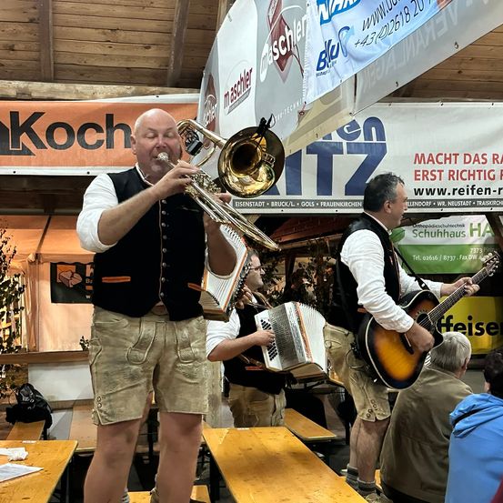 Ein Mann in traditioneller bayerischer Kleidung spielt Posaune, während ein anderer Akkordeon spielt. Ein dritter Mann spielt Gitarre vor einem Publikum.