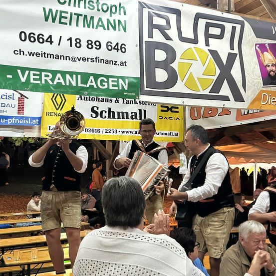 Vier Männer in traditioneller Kleidung spielen auf Instrumenten auf einem Freiluftfestival. Ein Banner mit dem Text 'RUPPI BOX' hängt im Hintergrund.