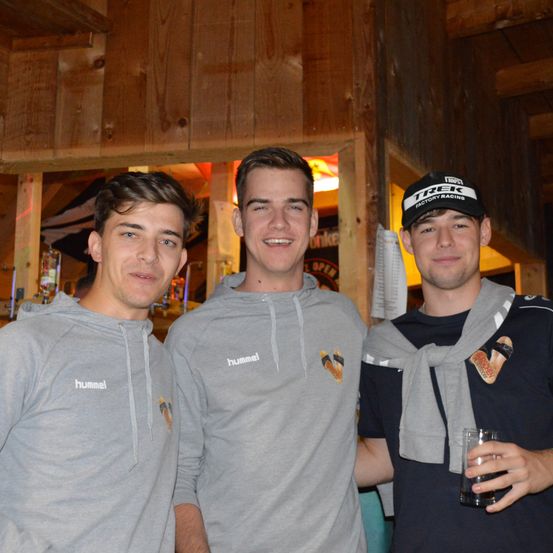 Drei Männer posieren drinnen, zwei tragen graue Hoodies mit 'hummel' vorne und einer eine schwarze Kappe mit 'TREK'. Der Mann rechts hält ein Glas. Dahinter eine Holzwand und eine Tür mit 'OFFEN' Schild.
