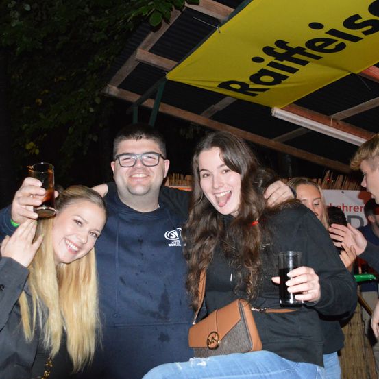 Drei junge Leute posieren für ein Foto bei einer nächtlichen Veranstaltung. Der Mann hat ein Bier in der Hand, während die beiden Frauen lächeln. Sie stehen unter einem Vordach mit dem Wort 'Raffeisen'.