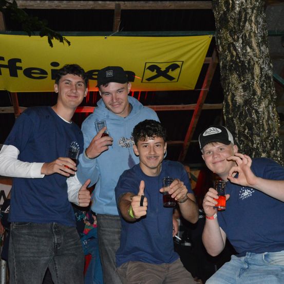 Vier junge Männer posieren nachts für ein Foto. Sie halten Getränke in den Händen und lächeln. Der Mann in der Mitte macht ein Friedenszeichen. Im Hintergrund ist ein gelbes Banner und ein Baum zu sehen.