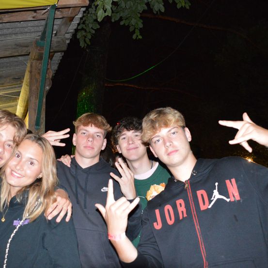 Fünf junge Erwachsene stehen bei Nacht unter einem Holzunterstand. Sie lächeln alle und machen Handzeichen. Der Mann in der Mitte trägt eine 'JORDAN'-Hoodie.