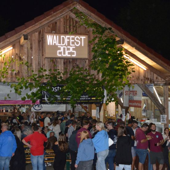 Eine Menschenmenge versammelt sich bei einer Waldfest-Veranstaltung im Jahr 2025 vor einem Holzgebäude mit einem Schild. Die Leute stehen und unterhalten sich, einige tragen Brillen.