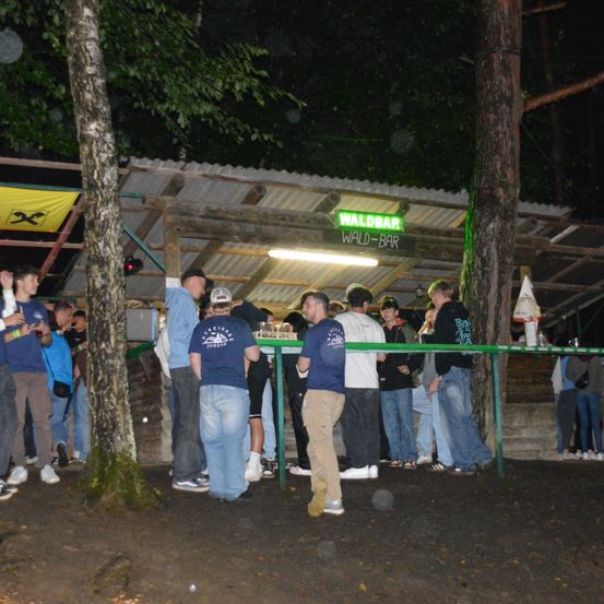 Eine Gruppe von Menschen steht bei Nacht in einer Schlange vor einer Bar. Ein Schild mit grünen Lichtern steht für Wald-Bar.