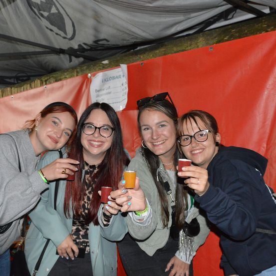 Vier Frauen lächeln und halten kleine Gläser, zwei rot, eines orange und eines blau, vor einem roten Hintergrund.
