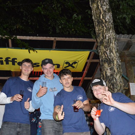 Fünf junge Männer stehen zusammen unter einem gelben Banner. Sie halten Getränke in den Händen, zwei von ihnen zeigen Daumen hoch, einer mit einem orangefarbenen Getränk. Ein Baumstamm steht hinter ihnen.