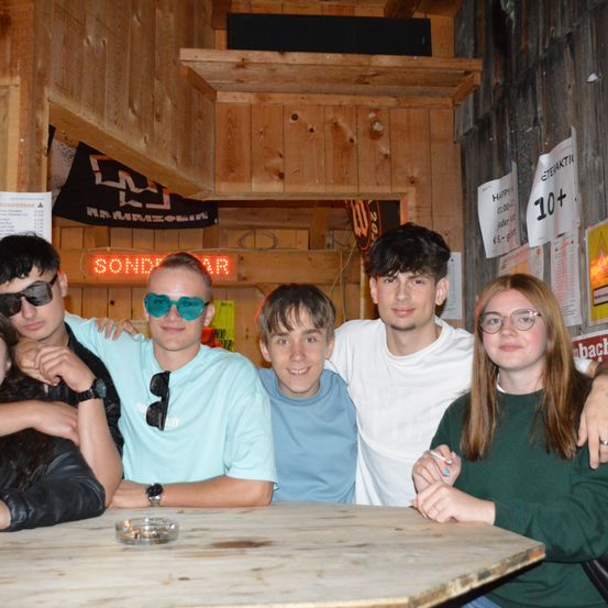 Sechs junge Leute sitzen an einem Holztisch und lächeln für ein Foto. Der Ort ist drinnen, mit Holzwänden und verschiedenen Plakaten. Ein Neonschild mit der Aufschrift 'Sonderbar' ist sichtbar.