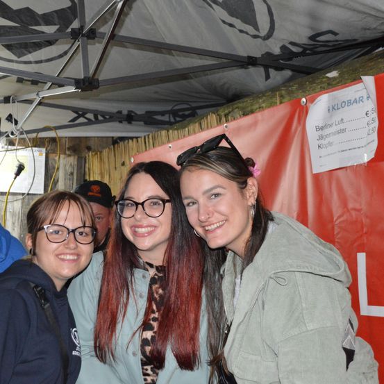 Drei Frauen lächeln und posieren für ein Foto unter einem Zelt mit rotem Hintergrund. Ein Schild mit Getränkepreisen ist an der Wand sichtbar.