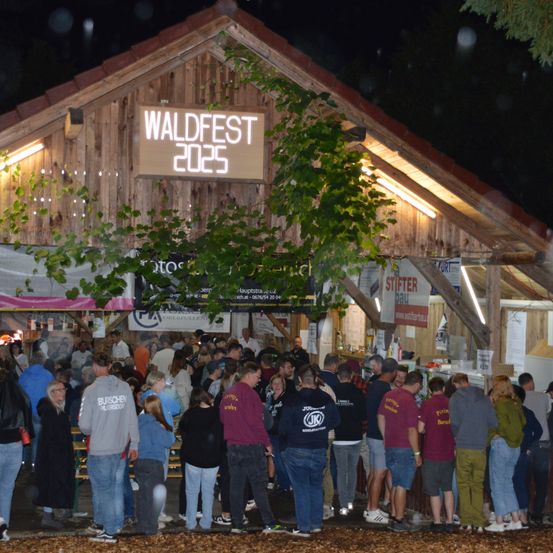 Eine Menschenmenge versammelt sich vor einem Gebäude mit einem Schild, das Waldfest 2025 anzeigt, während einer Nachtveranstaltung. Die Leute tragen Freizeitkleidung und einige halten Getränke. Das Gebäude hat Holzwände und ein Dach. Es gibt zusätzliche Schilder und Banner, die hängen.