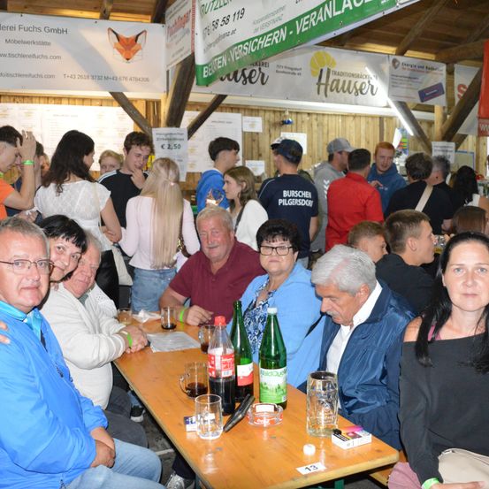Eine große Gruppe von Menschen ist auf einer Party versammelt. Sie sitzen um einen Tisch mit Getränken und Essen. Der Raum ist mit Bannern und Dekorationen gefüllt.