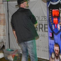 Ein Mann steht vor einer großen Spielmaschine, die Punkte anzeigt. Er trägt eine Mütze und eine Brille. Ein Plastikbehälter mit Gegenständen steht neben ihm. Ein Banner mit Text und Bildern ist hinter ihm.
