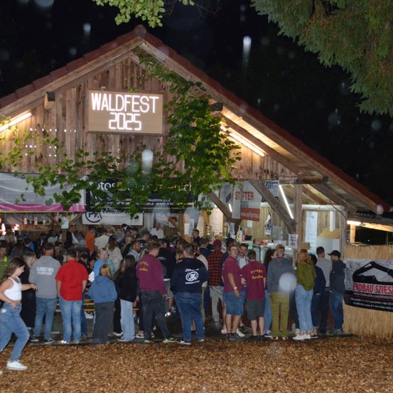 Eine nächtliche Zusammenkunft beim Waldfest 2025. Menschen stehen in einer Schlange vor einem Holzgebäude mit einem Schild darüber. Die Menge ist vielfältig und trägt verschiedene Kleidung. Einige haben Hüte auf, andere tragen Turnschuhe. Ein großer Baum steht neben dem Gebäude.